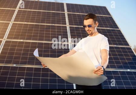 Junger Geschäftsmann schaut in einen Weißpapierplan, der neben einem großen Solarpanel steht, Sonne scheint hell auf dem Gesicht, umweltfreundliches Konzept Stockfoto