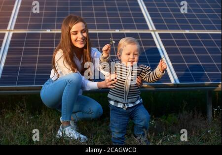 Junge Mutter unterstützt ihren kleinen Sohn bei seinen ersten Schritten in der Nähe von Sonnenkollektoren, Konzept der modernen Familie Stockfoto