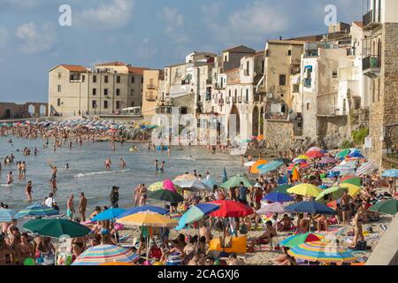 11.08.2018, Cefalu, Sizilien, Italien - Touristen und Einheimische entspannen sich am Stadtstrand unter Sonnenschirmen und baden im Mittelmeer. Im Hintergrund Stockfoto