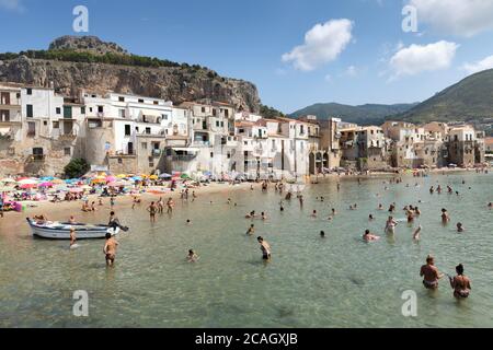 11.08.2018, Cefalu, Sizilien, Italien - Touristen und Einheimische entspannen sich am Strand unter Sonnenschirmen und schwimmen im Mittelmeer. Im Hintergrund Häuser von Stockfoto