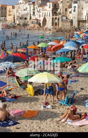 11.08.2018, Cefalu, Sizilien, Italien - Touristen und Einheimische entspannen sich am Stadtstrand unter Sonnenschirmen und baden im Mittelmeer. Im Hintergrund Stockfoto