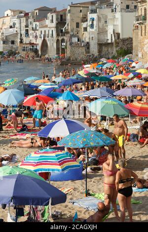 11.08.2018, Cefalu, Sizilien, Italien - Touristen und Einheimische entspannen sich am Stadtstrand unter Sonnenschirmen und baden im Mittelmeer. Im Hintergrund Stockfoto