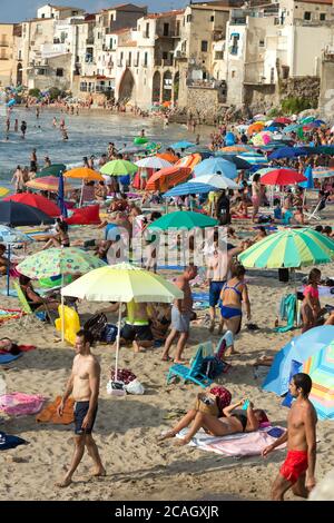 11.08.2018, Cefalu, Sizilien, Italien - Touristen und Einheimische entspannen sich am Stadtstrand unter Sonnenschirmen und baden im Mittelmeer. Im Hintergrund Stockfoto