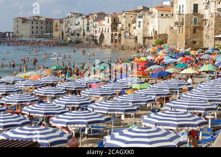 11.08.2018, Cefalu, Sizilien, Italien - Touristen und Einheimische entspannen sich am Stadtstrand unter Sonnenschirmen und baden im Mittelmeer. Im Hintergrund Stockfoto