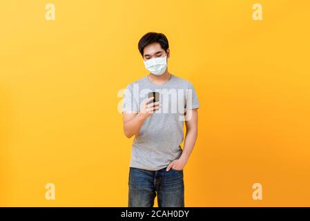 Junge asiatische Mann trägt medizinische Gesichtsmaske mit Handy Isoliert auf gelbem Studiohintergrund Stockfoto