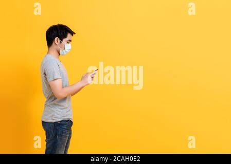 Junge asiatische Mann trägt medizinische Gesichtsmaske stehen und mit Mobiltelefon isoliert auf gelbem Studiohintergrund mit Kopierplatz Stockfoto