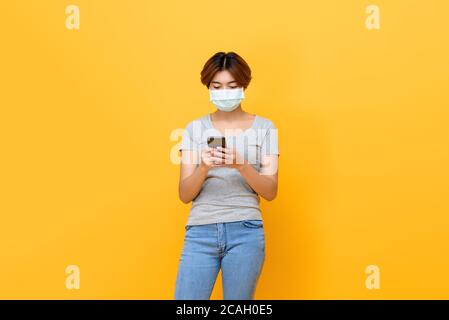 Junge asiatische Frau trägt medizinische Gesichtsmaske mit Handy Isoliert auf gelbem Studiohintergrund Stockfoto