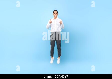 Glücklich lächelnd energisch asiatischen Mann in semi formale Kleidung springen In der Luft mit zwei Händen, die isoliert nach oben zeigen Hellblauer Hintergrund Stockfoto