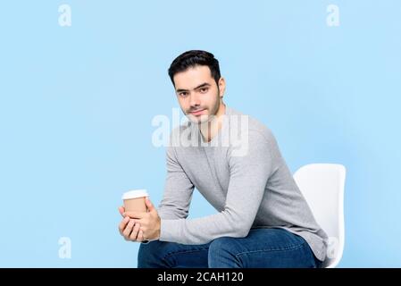 Porträt eines lächelnden jungen gutaussehenden kaukasischen Mann Blick auf die Kamera Während Sie sitzen und halten Kaffeetasse isoliert auf hellblau studio-Hintergrund Stockfoto