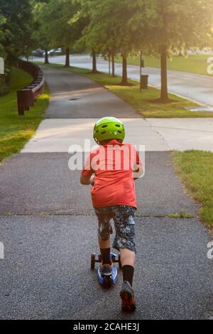 Bild eines Kindes mit grünem Helm, orangefarbenem T-Shirt und capri-Hosen, das auf einem Radweg in einem Vorort fährt. Es gibt Bäume Stockfoto