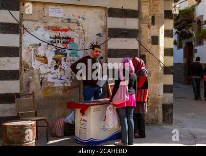 Damaskus, Syrien 03/29/2010: Ein junger, gut aussehender Verkäufer der syrischen Straße versucht, Ornamente an zwei arabische Frauen zu verkaufen. Er zeigt Elemente auf Notbehelfsbehelf an Stockfoto