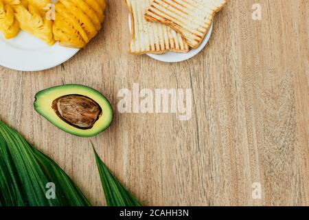 Flat Lay von gesunden Frühstück aus Obst und Toast. Geschälte Ananas auf einem Teller und ein gesundes Avacado für ein Toast-Sandwich auf einem Holztisch. Co Stockfoto