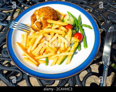 Ein Mittagessen mit geräuchertem Schellfisch-Kuchen mit französischen Bratkartoffeln, Spargeltomaten und Gurken, serviert im Freien auf einem Außentisch Stockfoto