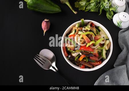 Salat mit Auberginen, Tomaten, Paprika und Gurken in weißer Schüssel auf schwarzem Hintergrund, Platz für Text Stockfoto