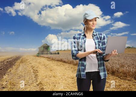 Junge Landwirtschaftsingenieurin mit Laptop und Mähdrescher, die in einem Weizenfeld arbeiten Stockfoto