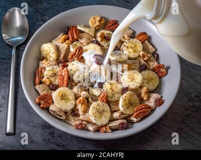 Nahaufnahme POV-Aufnahme von Milch, die aus einem Karton über Vollkorngetreide, Bananenscheiben, Pekannüsse und Walnüsse, Rosinen und Chiasamen gegossen wird. Stockfoto