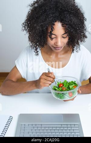 Schöne junge schwarze Frau essen einen Salat vor Ein moderner Laptop mit weißem Hintergrund Stockfoto