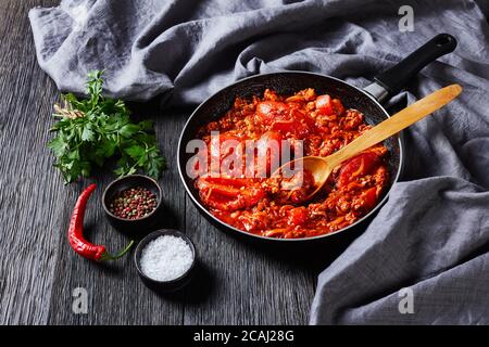 Ragout mit ganzen Pflaumentomaten, Zwiebelscheiben, gemahlenen italienischen Schweinewürsten und Gewürzen in einer Pfanne auf einem dunklen Eichenholztisch mit grauem Tuch, Mok Stockfoto