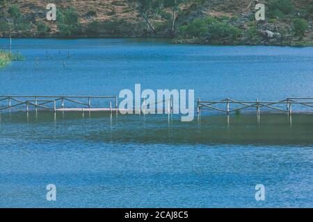 Alte teilweise versunkene Holzbrücke über einen ruhigen See umgeben Von Bäumen und Sträuchern Stockfoto