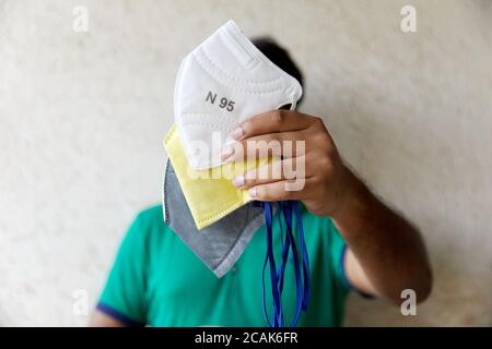 Der indische Mann hält drei bunte Atemschutzmasken vor sich Kamera gegen eine beige strukturierte Wand Stockfoto