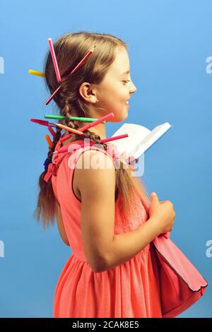 Zurück zur Schule Konzept. Schulmädchen mit glücklich lächelnd Gesicht isoliert auf blauem Hintergrund. Kind in rosa Kleid und Schultasche trägt Zöpfe mit Buntstiften. Mädchen hält Tasche und offenes Notebook, Seitenansicht Stockfoto