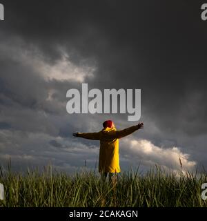 Mann in gelber Regenjacke und rotem Hut mit weit geöffneten Händen, der bei Regenwetter auf dem Feld steht, blickt auf den dramatischen bewölkten Himmel. Herbstsaison. Stockfoto