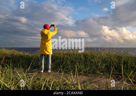 Mann in gelber Regenjacke tragen roten Hut stehen am Strand am Holzweg, schaut auf dramatische bewölkten Himmel und macht Foto auf dem Smartphone. Herbstsaison. Stockfoto