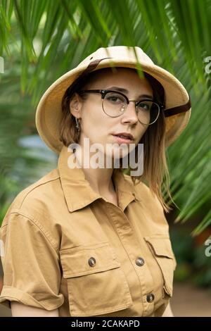 Frau Botanikerin im Safaristil im Gewächshaus gekleidet. Naturforscher in Khaki-Kleidung, Lever Handschuhe im Regenwald von Palmen umgeben, Blick auf die Kamera. Stockfoto