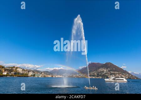 Schöne Panoramasicht auf Lugano See und Stadt mit Berg Monte Bre und Schweizer Alpen im Hintergrund Brunnen und Regenbogen, Kreuzfahrtschiff im Vordergrund, C Stockfoto