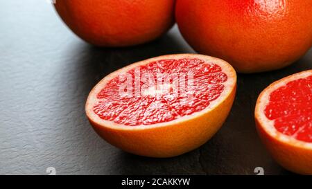 Nahaufnahme - Hälfte der rosa Grapefruit auf dunklem Arbeitsbrett, mehr Zitrusfrüchte im Hintergrund Stockfoto