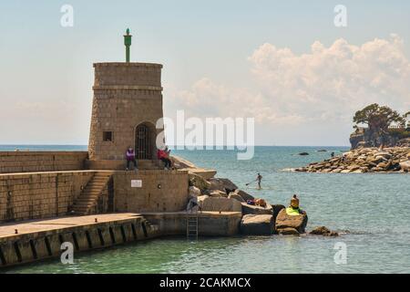 Ein steinerner Leuchtturm mit Blick auf den Golf des Paradieses mit Menschen sitzen auf den Felsen und ein Mann auf einem Stand up Paddel im Sommer, Recco, Genua, Ligurien Stockfoto