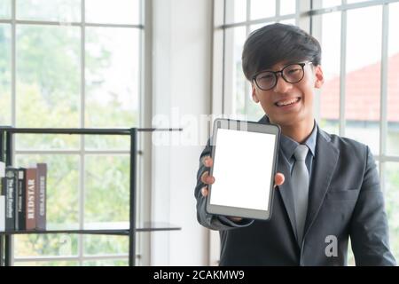 Junge asiatische Geschäftsmann trägt einen Anzug halten und zeigt Vorderansicht einer weißen leeren Bildschirm Tablet mockup. Konzept der professionellen stellen Sie sich vor Stockfoto