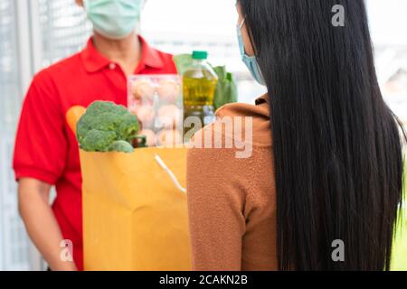 Asiatische Lieferung Mann aus dem Supermarkt trägt eine Gesichtsmaske und hält eine Tasche mit frischen Lebensmitteln, Gemüse und Obst für die Kunden zu Hause geben. Conce Stockfoto