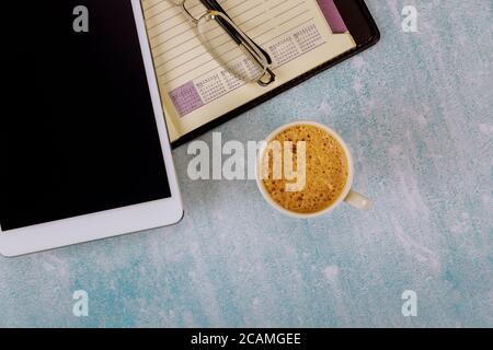 Ein offenes wöchentliches Notizbuch und digitale Tablet-Brillen auf dem Tisch eine weiße Tasse Kaffee. Stockfoto