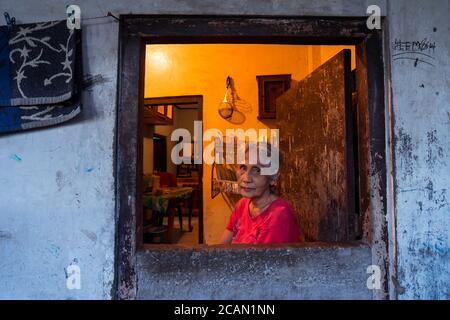 Yogyakarta / Indonesien - 2. August 2018: Porträt einer alten indonesierin mit weißen Haaren, die durch die Öffnung vor der Haustür auf die Kamera schaut Stockfoto