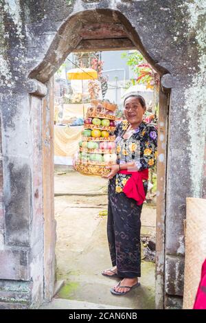 Bali / Indonesien - 15. August 2018: Portrait einer buddhistischen Frau, die vor der Tür eines kleinen lokalen Tempels lächelt und Früchte als religiöse Opfergabe hält Stockfoto