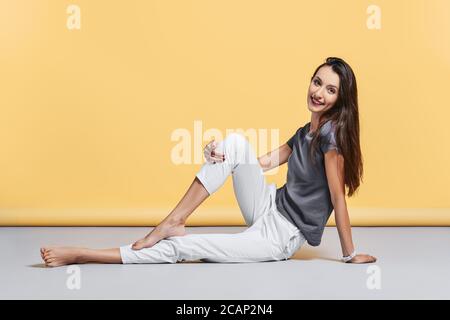 Happy barfuß junge Frau in lässigen Hosen und Hemd sitzt auf dem Boden im Studio auf gelbem Hintergrund Stockfoto