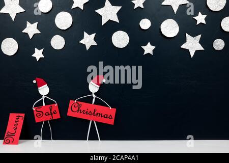 Stick Mann Figur trägt santa Hut hält eine sichere Weihnachten Grußkarte mit silbernen Sternen und Kugeln Ornament Hintergrund. Covid Weihnachtsfeier Konzept. Stockfoto