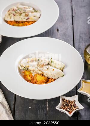 Zwei Portionen Heilbutt-Filet gebacken mit Gemüse und Kartoffel-Confit garnieren in einem weißen Teller. Restaurantrezepte. Köstliche natürliche Meeresfrüchte. Vertikale Stockfoto