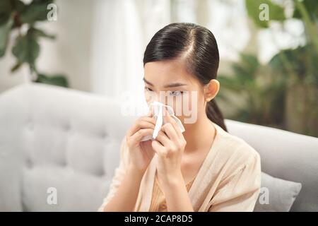 Kranke junge Frau sitzt auf dem Sofa, bläst ihre Nase zu Hause im Wohnzimmer. Foto von niesen Frau in Papiergewebe. Stockfoto