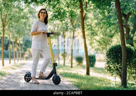 Fröhliche liebenswert ältere Frau mit einem Roller während der Fahrt im Park. Moderne Frau, eine neue Generation. Gesunde, fröhliche ältere Dame im Ruhestand. Konzept Stockfoto