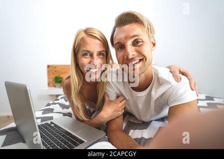 Junges Paar unter Selfie im Bett - glücklich Liebhaber mit Spaß beim Fotografieren selbst auf dem Bett liegend, während mit Laptop Im Schlafzimmer Stockfoto