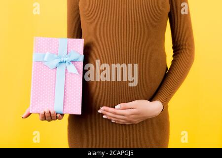 Zugeschnittenes Bild von rosa gefleckte Geschenkbox in der Hand der schwangeren Frau gegen ihren Bauch auf gelbem Hintergrund. Zukünftige Mutter in braunem Kleid. Warten auf einen BA Stockfoto