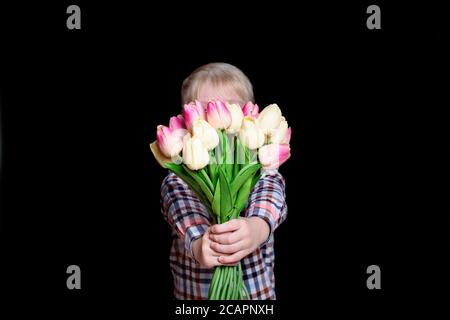 Der kleine Junge versteckt sein Gesicht hinter einem Blumenstrauß aus Tulpen. Auf schwarzem Hintergrund isolieren. Stockfoto