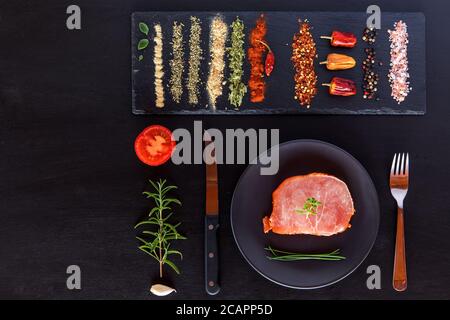 Draufsicht rohes Schweinekotelett Steak auf schwarzem Holzhintergrund. Stücke von rohem Schweinesteak mit Gewürzen und Kräutern Rosmarin, Thymian, Basilikum, Salz und Pfeffer Stockfoto