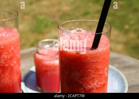 Nahaufnahme von isolierten eiskalten Wassermelone Saft Glas mit Stroh draußen auf dem Tisch. Scheiben von roten Früchten auf dem Teller. Stockfoto
