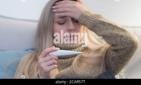 Nette junge kaukasische blonde Frau in Pullover liegt im Bett unter der Decke, Kopfschmerzen und Blick auf Thermometer, berühren ihre Stirn, wählen Stockfoto