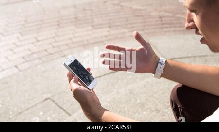 Mann hält gebrochenes Handy nach dem Tropfen, Blick von oben, im Freien. Smartphone mit kaputter (rissiger) Anzeige. Abgebrochenes Telefon aus dem f Stockfoto