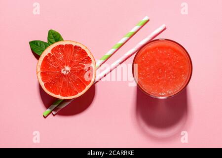 Glas frisch gepresster Grapefruitsaft mit Fruchtfleisch, halbe Grapefruit und zwei Trinkhalme werfen Schatten auf einen rosa Hintergrund. Aufnahme mit hartem Licht. Stockfoto
