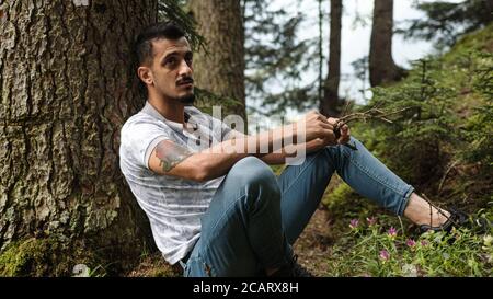 Hübscher junger Mann, der sich auf Baum stützt, beschäftigt sich mit Holzstäbchen in der Hand in Nahaufnahme, Entspannungskonzept Stockfoto